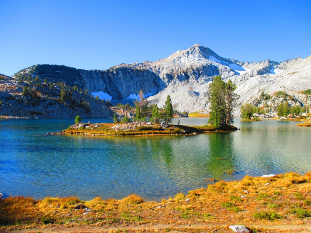 View of a pristine, blue alpine lake with a small island, featuring a few pine trees. Across the lake, on the opposite shore, are mountain ridge cliffs and snow-covered mountain side and summit of Glacier Peak.