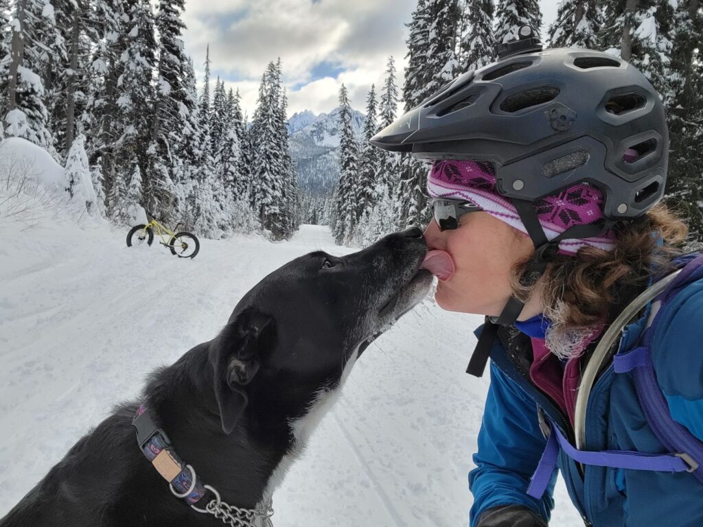 Dog with black fur licking the owner's face along a snowy mountain forest trail. Owner is wearing a bike helmet, and in the background is a fat bike.