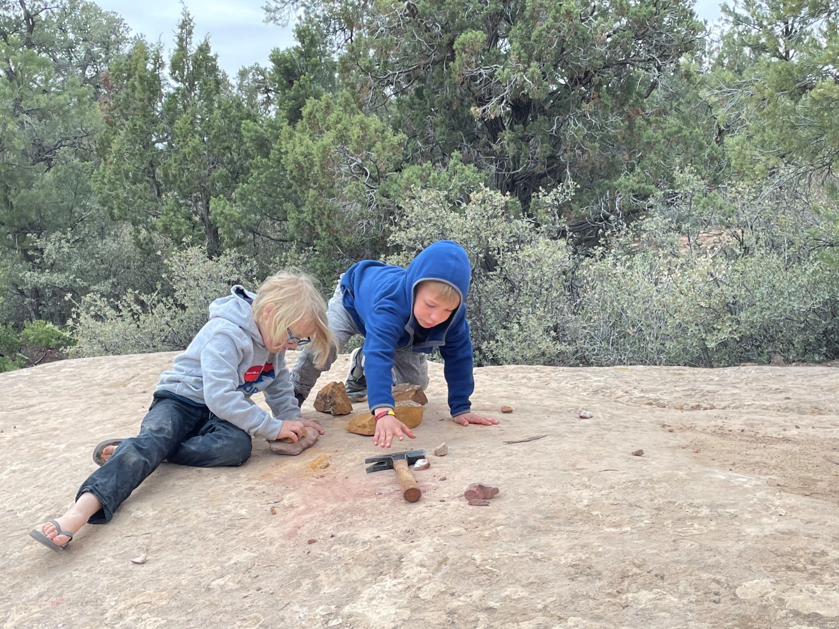 Two children laying down on a huge boulder examining a collection of rocks, with a hammer to break open rocks and explore the insides.on of small rocks and examining them.