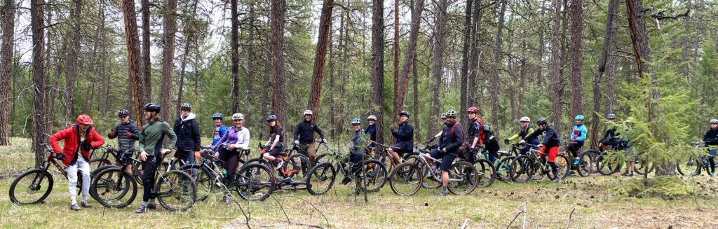 A long line of teenagers on their mountain bikes along a forested trail, stopped and smiling at the camera.