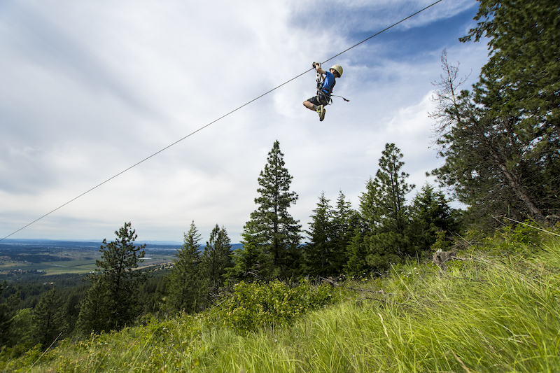 Child ziplining down a cable, smiling at the camera, over a grassy hillside with pine trees in the background.