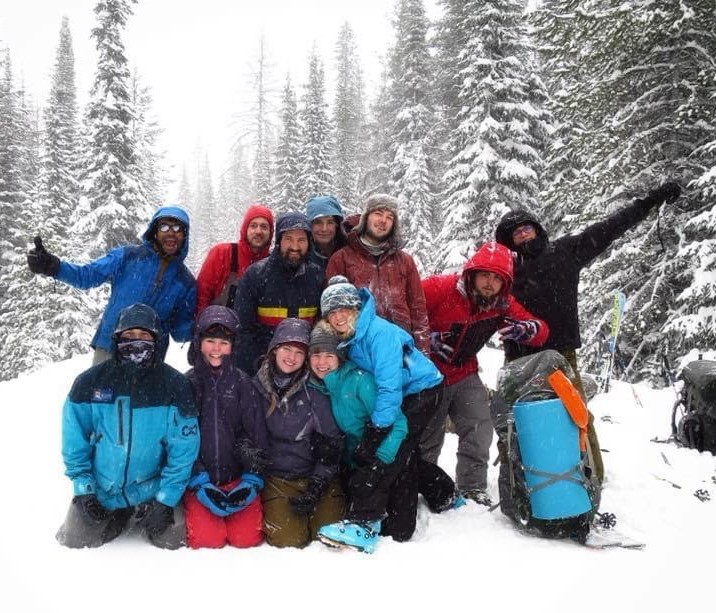 Group of 10 female and male university students dress in outdoor winter-expedition gear and smiling at the camera while standing and kneeling in the snow.