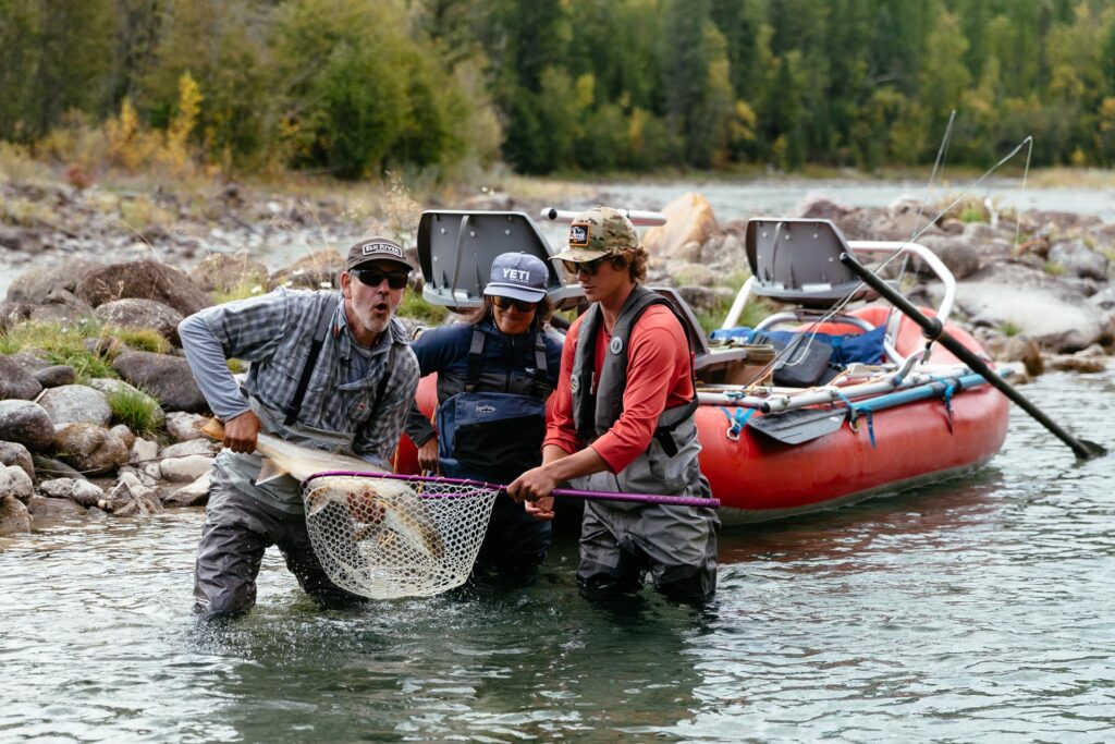 Three people holding a fishing net and one of them holding a large native trout while wade-standing in the Elk River in Canada, with a fishing raft behind them.
