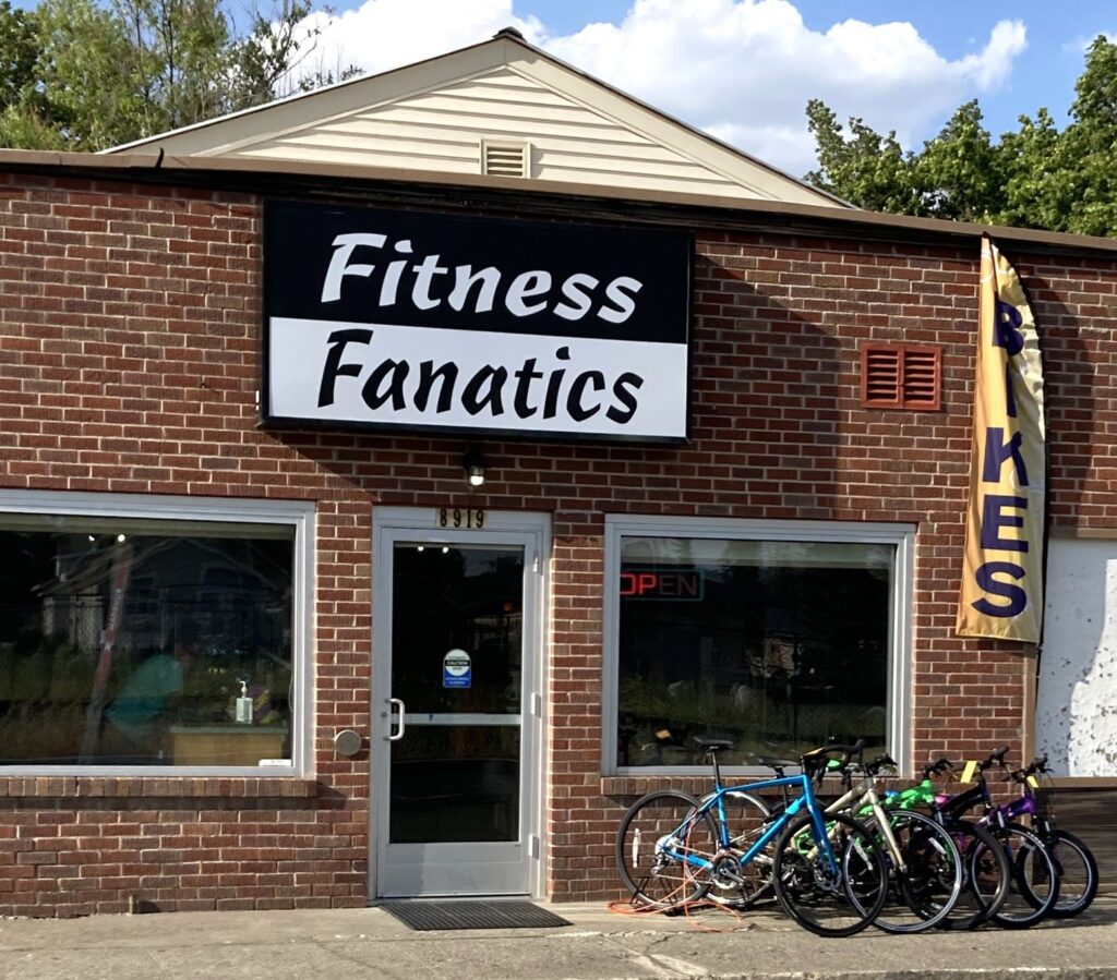 Storefront for Fitness Fanatics as its new location in Millwood, Wash., with brick wall, two glass windows, glass front door, bike rack out front, and large black and white store name sign.