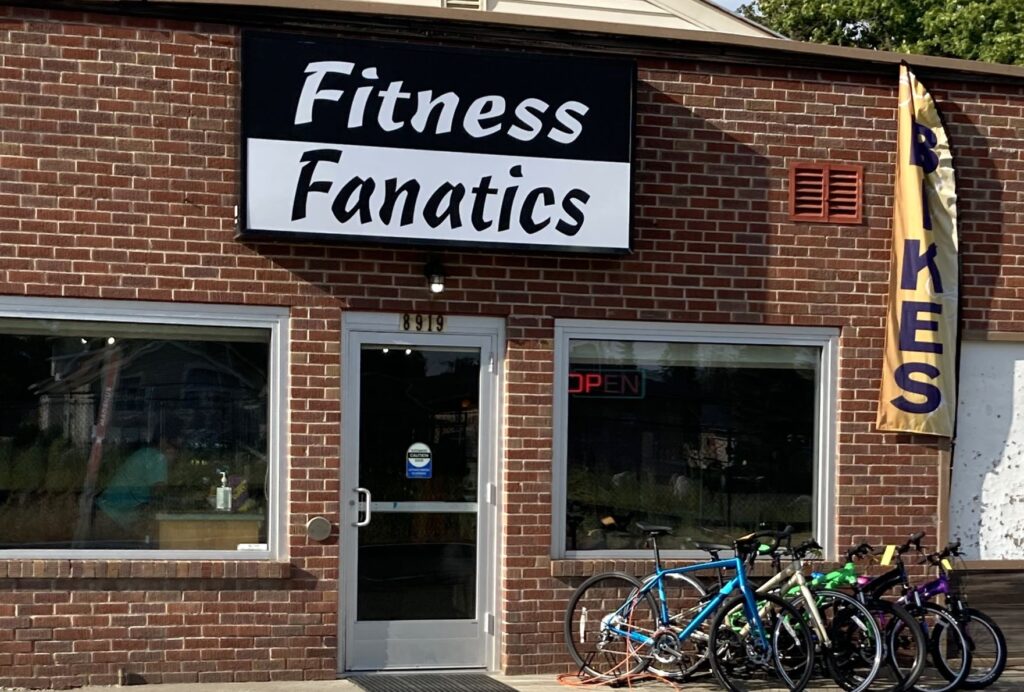 Storefront for Fitness Fanatics as its new location in Millwood, Wash., with brick wall, two glass windows, glass front door, bike rack out front, and large black and white store name sign.