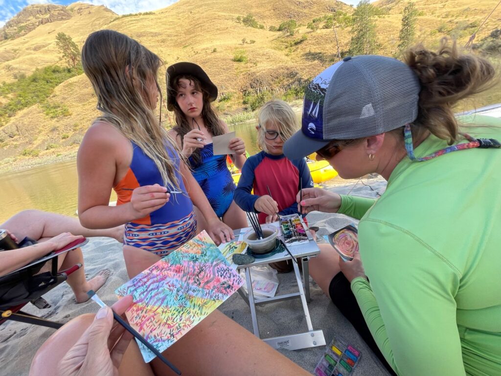 Adventure travel-river rafting and camping: Children and parents watercolor painting on a sandy beach during a river rafting and camping trip.