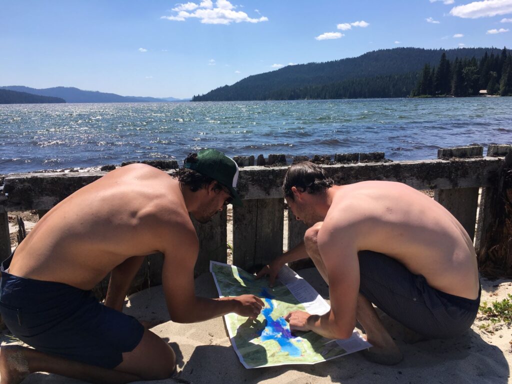 Two men squatting to examine a nautical map of Priest Lake, with view of the lake in the distance.