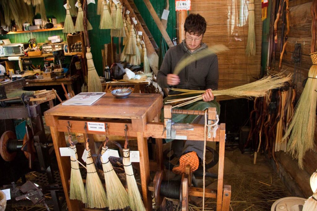 Man standing at a table, weaving natural materials together to create a broom, at the North Woven Broom Co.