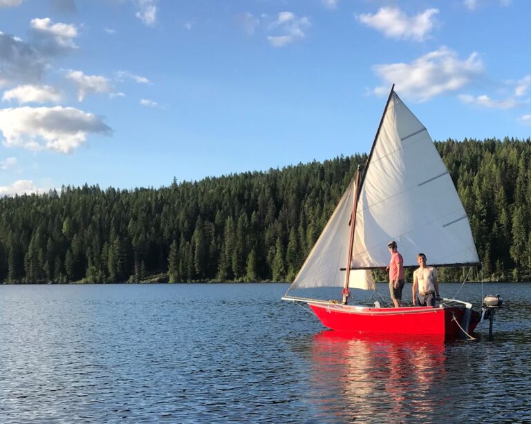 Author and his friend standing in a red sailboat on Upper Priest Lake with white sales open.