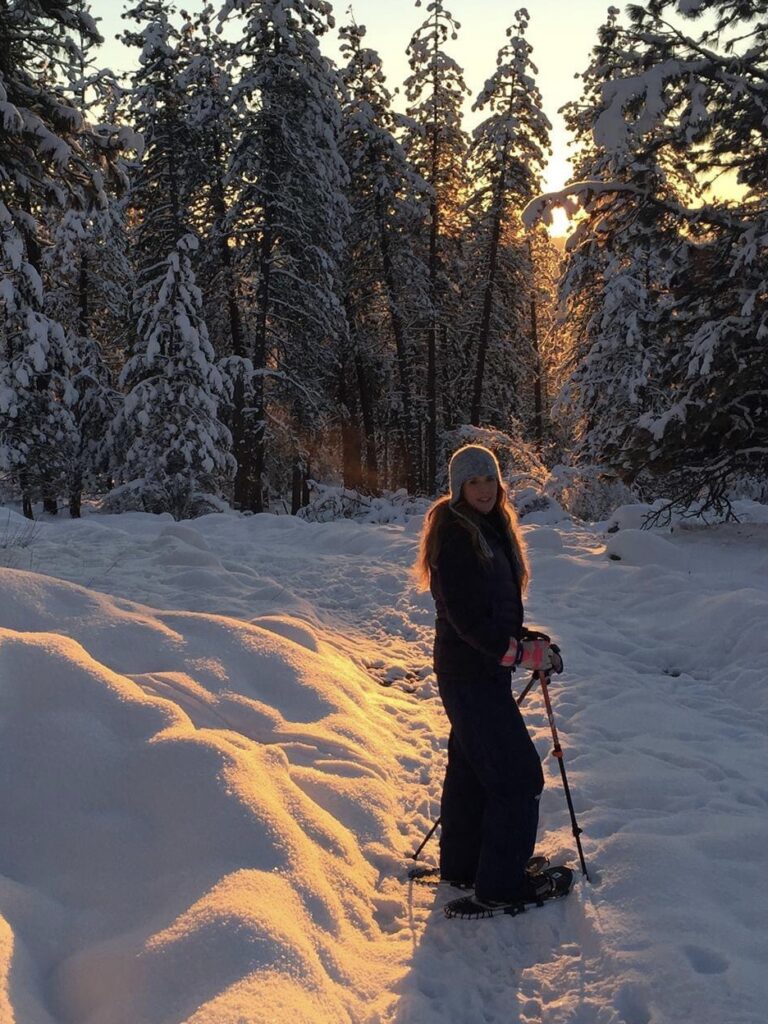 Woman snowshoeing with sun shining behind treetops in the background.