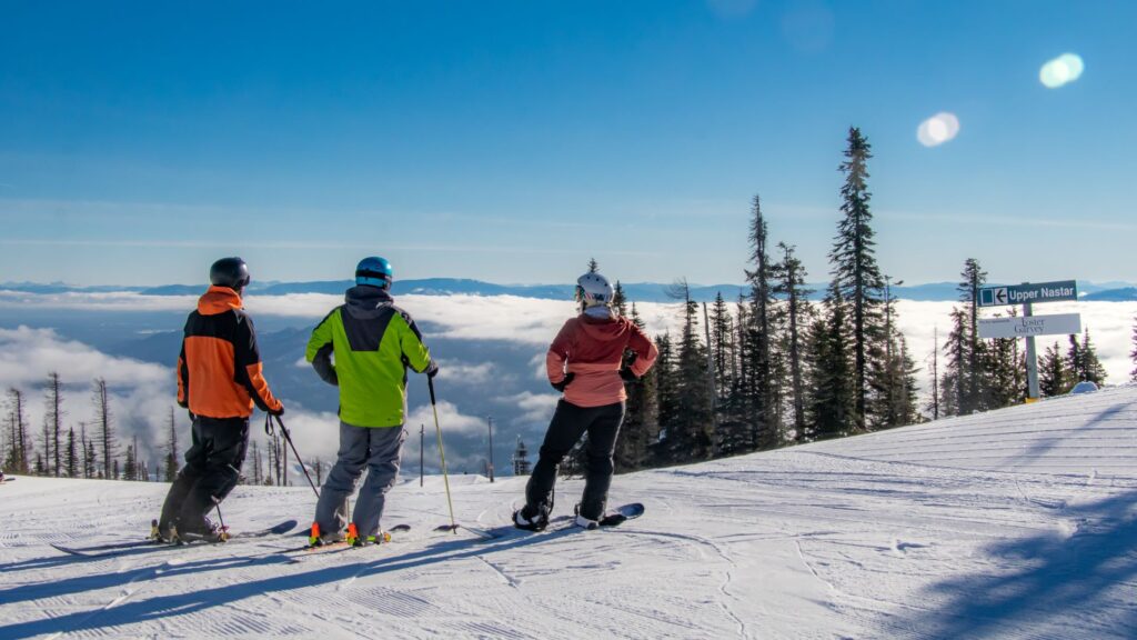Two skiers and a snowboarder at the summit of Mt. Spokane on a sunny, blue-sky day, looking at the view before going down a black diamond run.