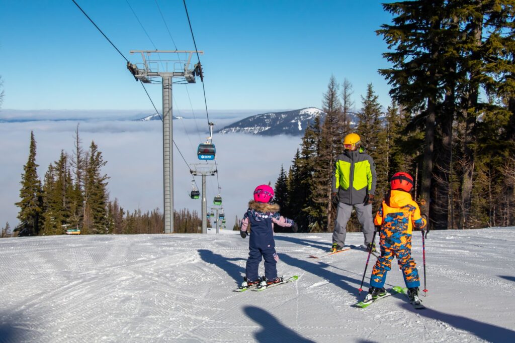 Two children on skis with an adult looking on as they ready to go down a slope on a sunny, blue sky day.